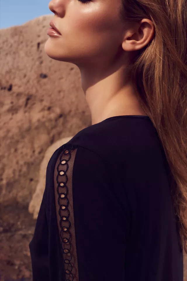 The image features a close-up profile of a woman with long, wavy hair. She is wearing a black top with a unique design on the sleeves, which includes a mesh segment with circular cutouts. The background consists of textured rock formations, suggesting a natural outdoor setting. The lighting accentuates her features, highlighting her skin and creating a serene atmosphere.