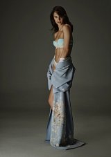 The image features a model standing gracefully against a dark background. She is wearing a light-colored, strapless bra and has a floral-embroidered shawl draped around her. The shawl is a soft blue, with intricate designs that enhance the visual appeal. The model strikes a poised pose, showcasing confidence. The overall mood of the image is elegant and artistic, with a focus on fashion and form.